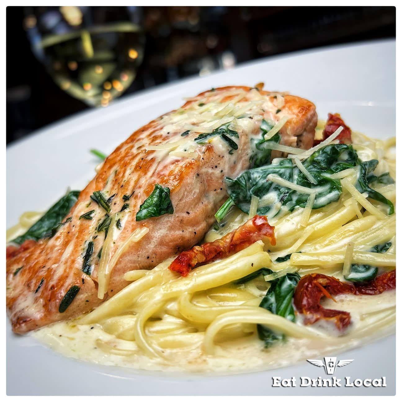 Salmon fillet with creamy sauce, spinach, and sun-dried tomatoes over pasta. White wine in the background.