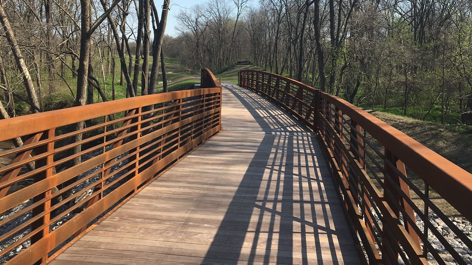 A wooden pathway bridge with rusty metal railings over a small ravine, surrounded by trees.