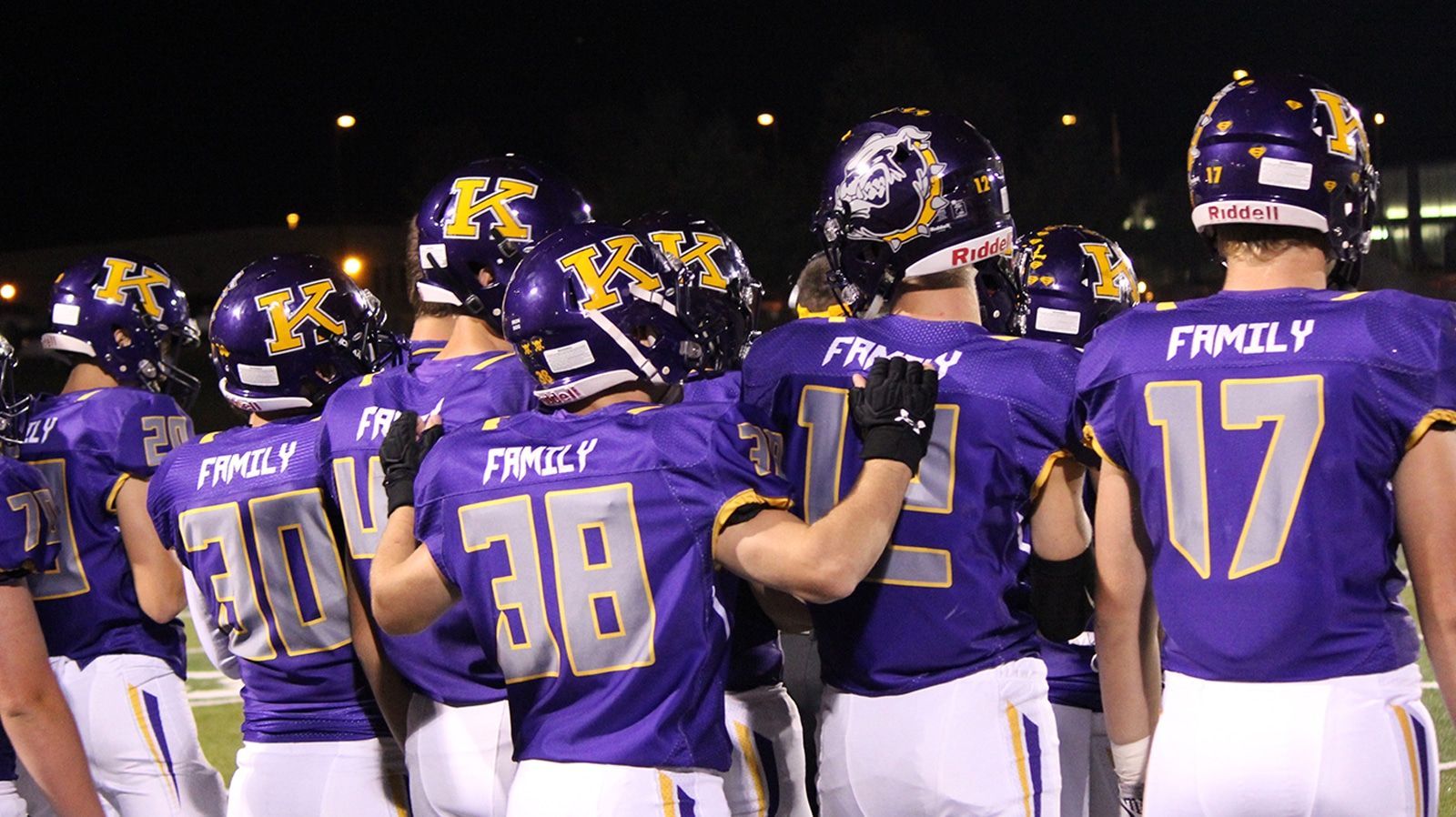 Football players in purple and gold uniforms huddle on a field at night. 