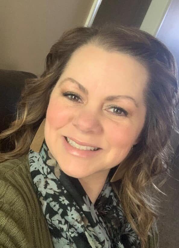 Woman with brown wavy hair and rosy cheeks smiles at the camera, wearing a green sweater and patterned scarf.