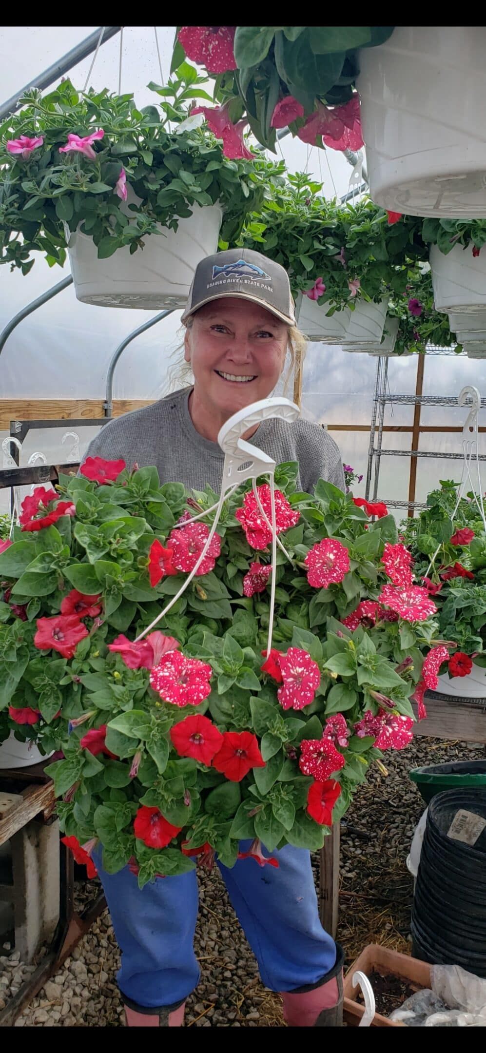 Woman in a cap and gloves smiles, holding a large hanging basket overflowing with red and pink flowers in a greenhouse.