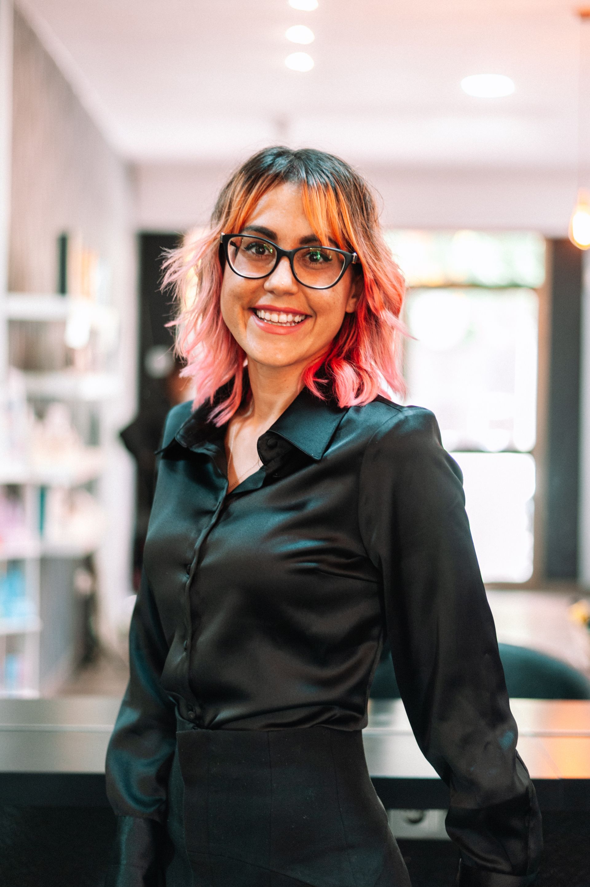 Woman with pink-streaked hair smiles, wearing glasses and a black satin blouse. She poses inside a brightly lit salon.