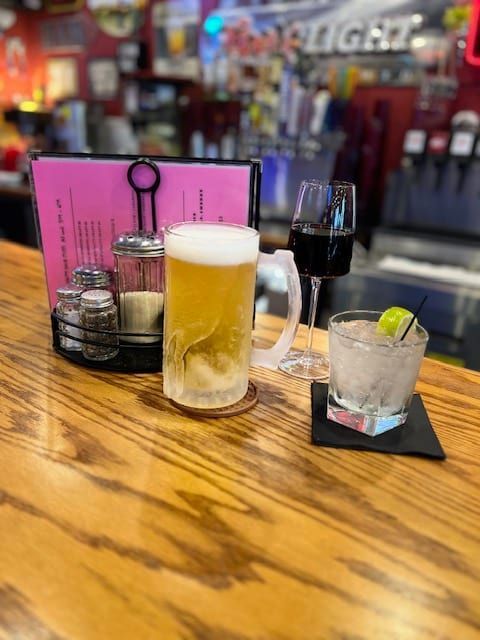 A bar with a frosty beer mug, a glass of red wine, and a cocktail on a wooden counter.