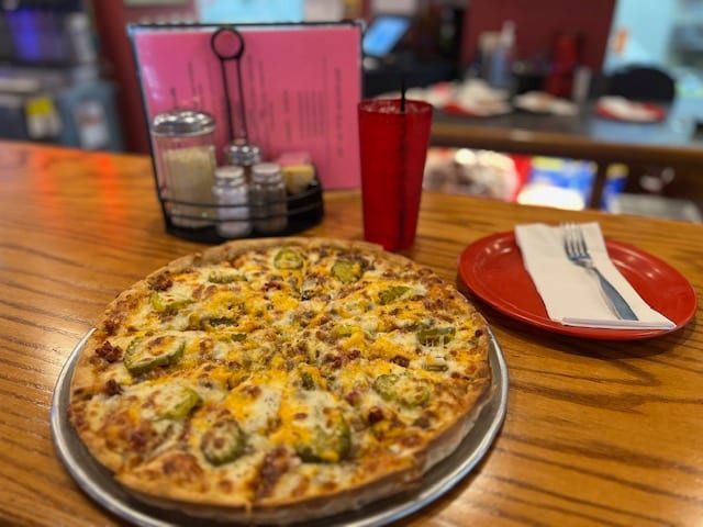 A jalapeño pizza on a metal tray, red drink, and plate with a fork on a wooden bar. Salt and pepper shakers and a pink menu are in the background.