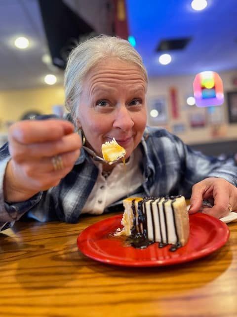 Woman eating cheesecake in a restaurant, gazing at the camera. The dessert is on a red plate, with a brown wood table.