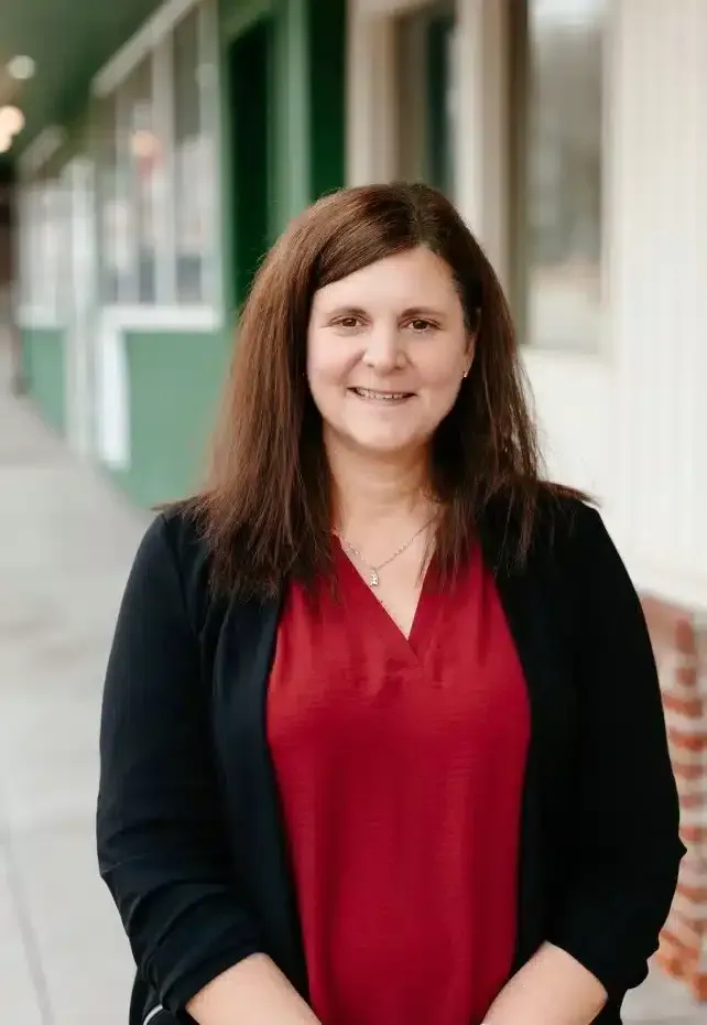 Woman with brown hair, wearing a red blouse and black cardigan, smiling outdoors in front of a green and white building.