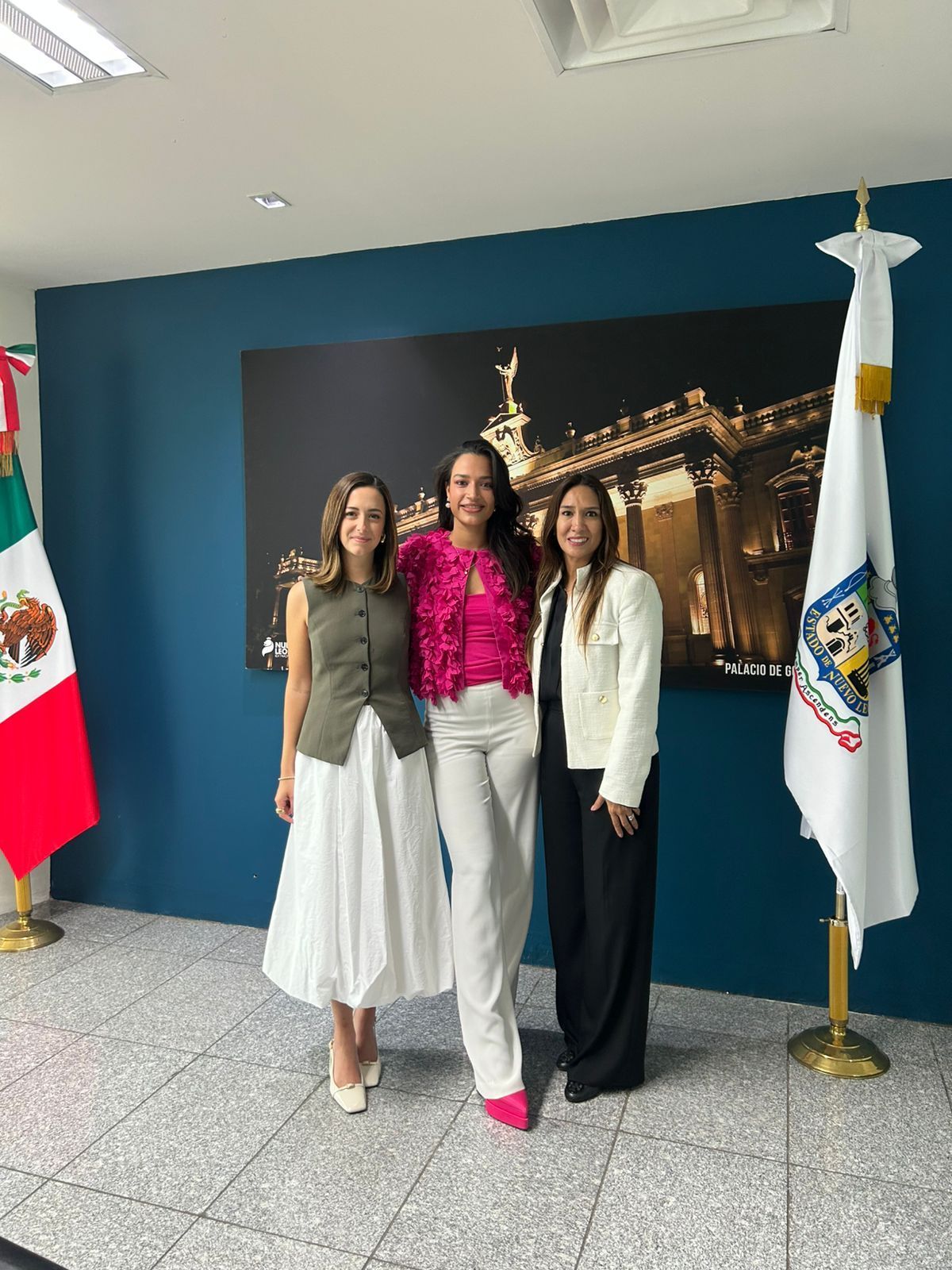 Tres mujeres posan en una oficina. Están de pie frente a una pared azul y la fotografía de un edificio; banderas a ambos lados.