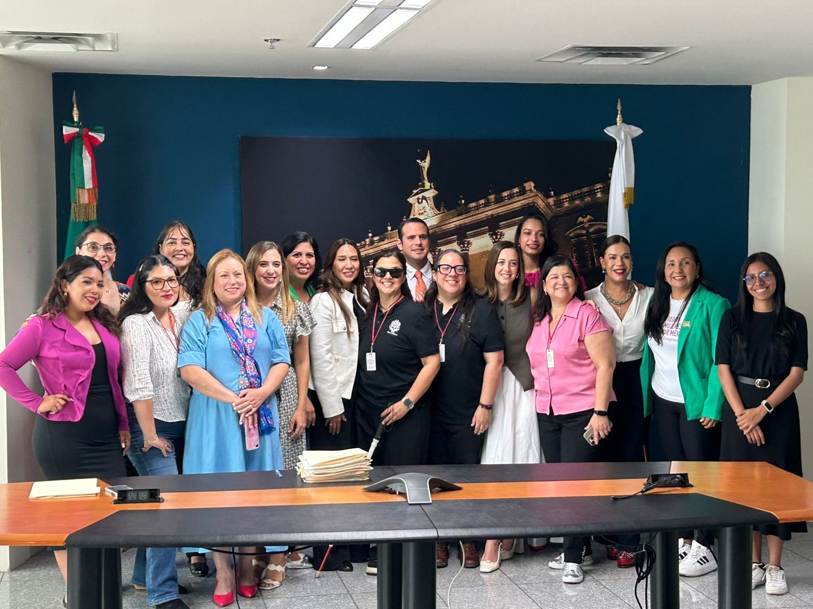 Grupo de personas posando para una foto frente a una pared azul con un fondo de ciudad y banderas.
