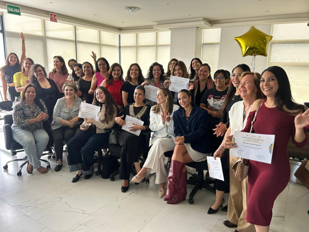 Grupo de mujeres en la oficina, con certificados en la mano, sonriendo a la cámara. Un globo dorado con forma de estrella flota.