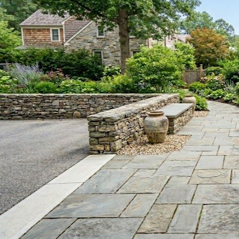 A stone patio with a low dry-stack stone wall, a ceramic urn, and a bench, leading toward a house and lush garden.