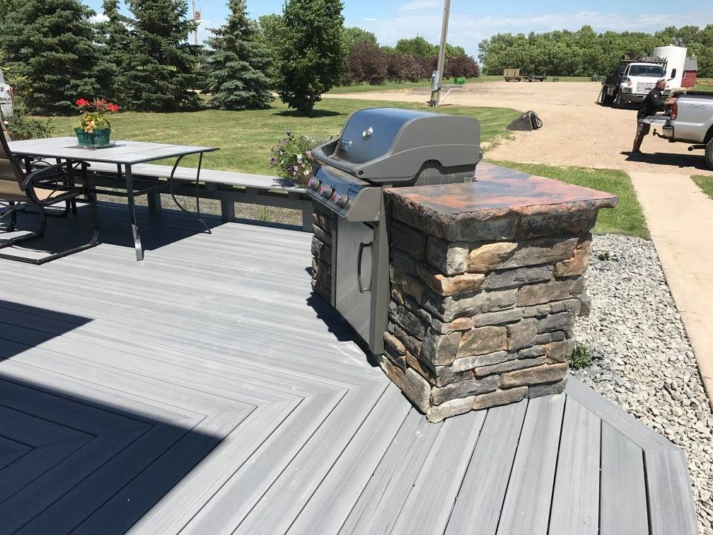 Outdoor grill built into a stone structure on a gray deck next to a table and chairs, on a sunny day.