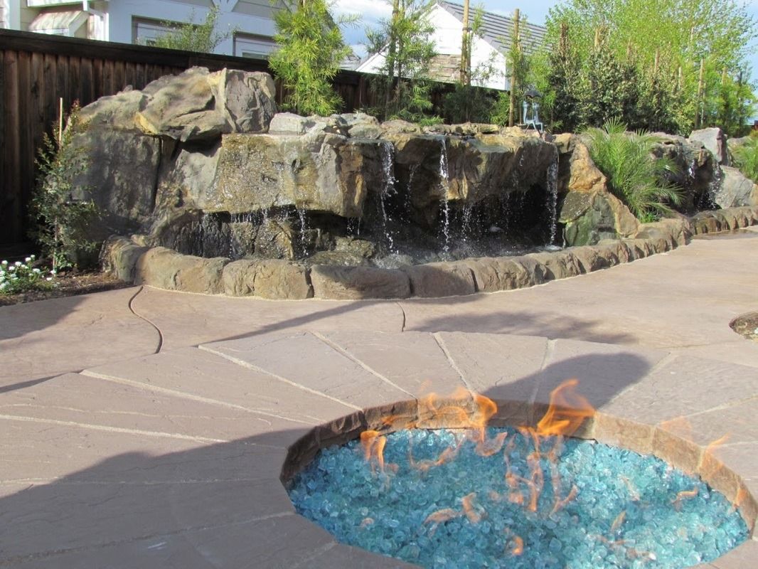 Water feature with waterfall and fire pit. Brown rocks, blue fire, stone patio, wooden fence.