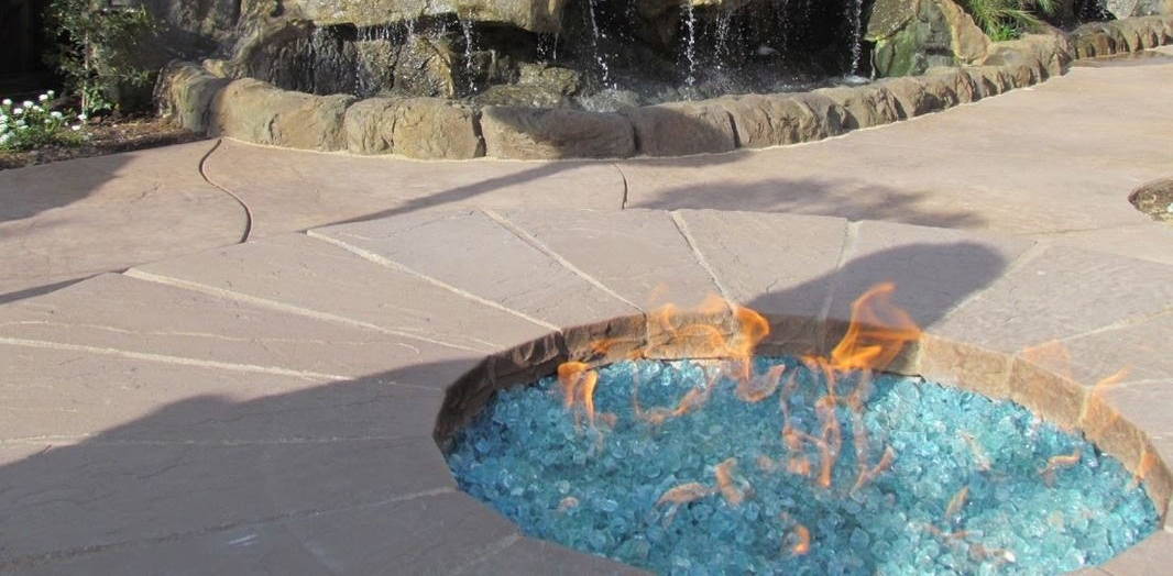 Fire pit with blue glass, flames, and stone patio near a waterfall.