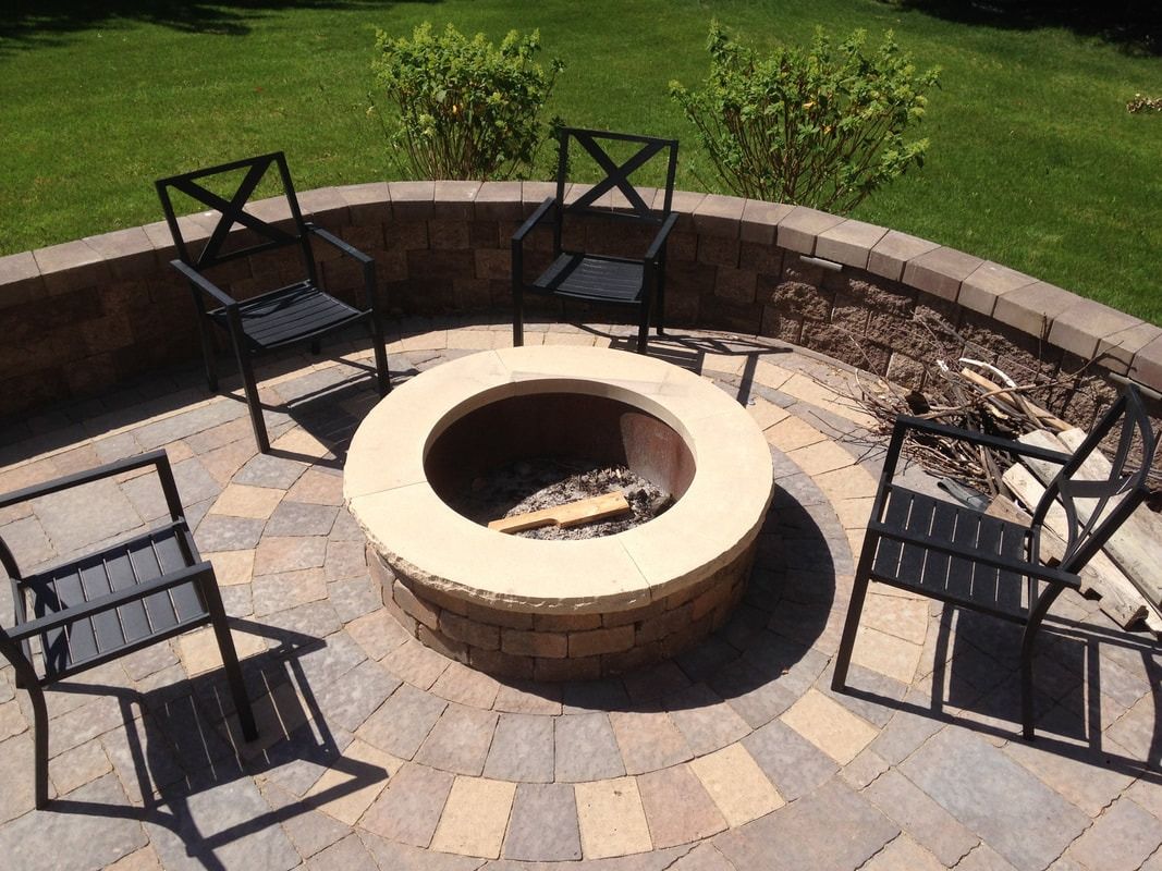 Stone fire pit with four black chairs on a circular patio, surrounded by a brick wall and green grass.