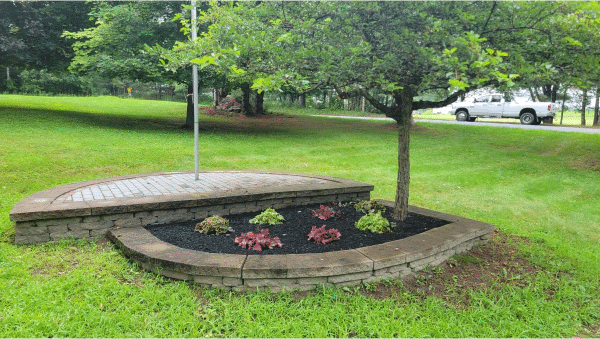 Stone-bordered flower bed with small tree, mulch, and colorful plants on green lawn. A white truck passes by a nearby road.