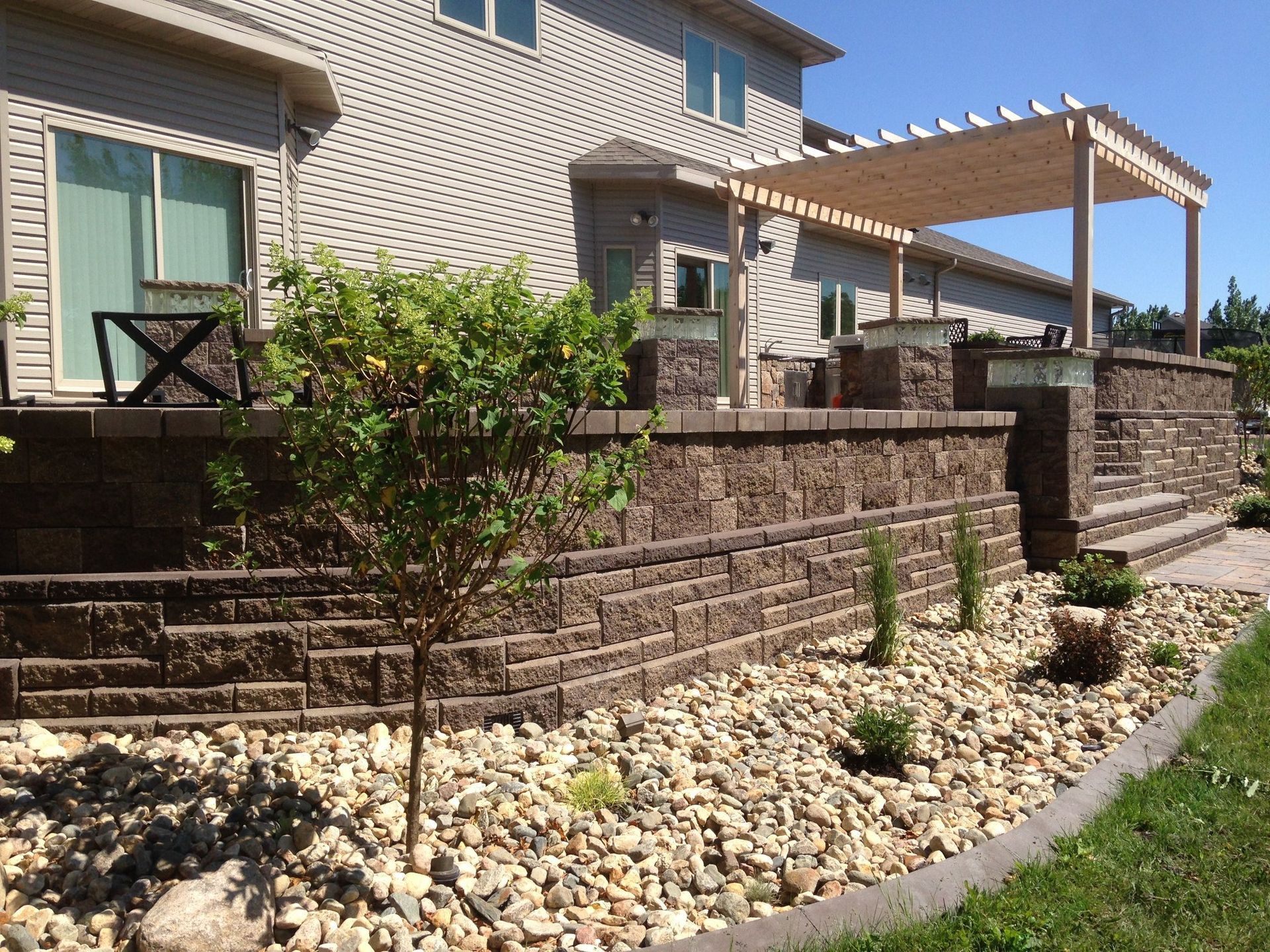 A gray house with a small tree in front, framed by wood and mulch, on a sunny day.