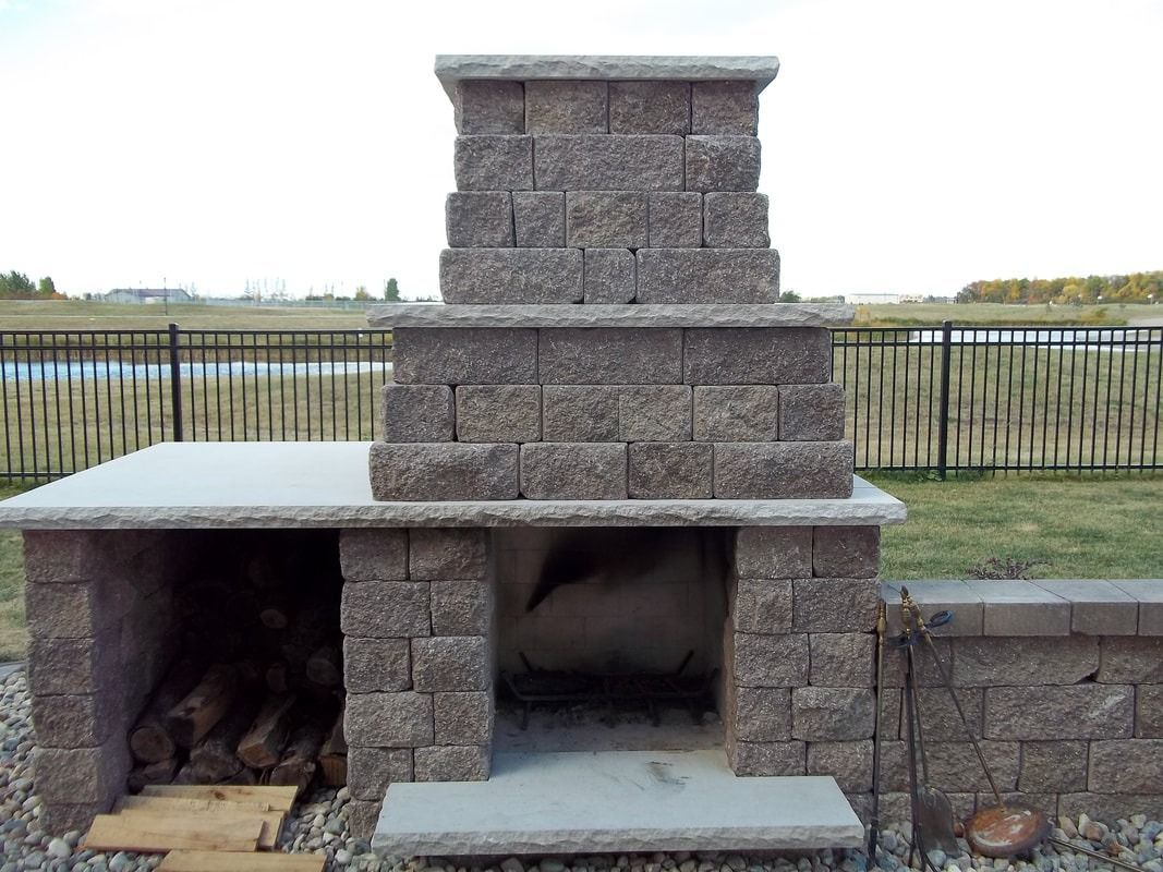Outdoor stone fireplace with a wood storage area on a grassy lawn near a black fence.