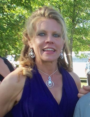 Woman with blonde hair, blue dress, and jewelry smiling outdoors.