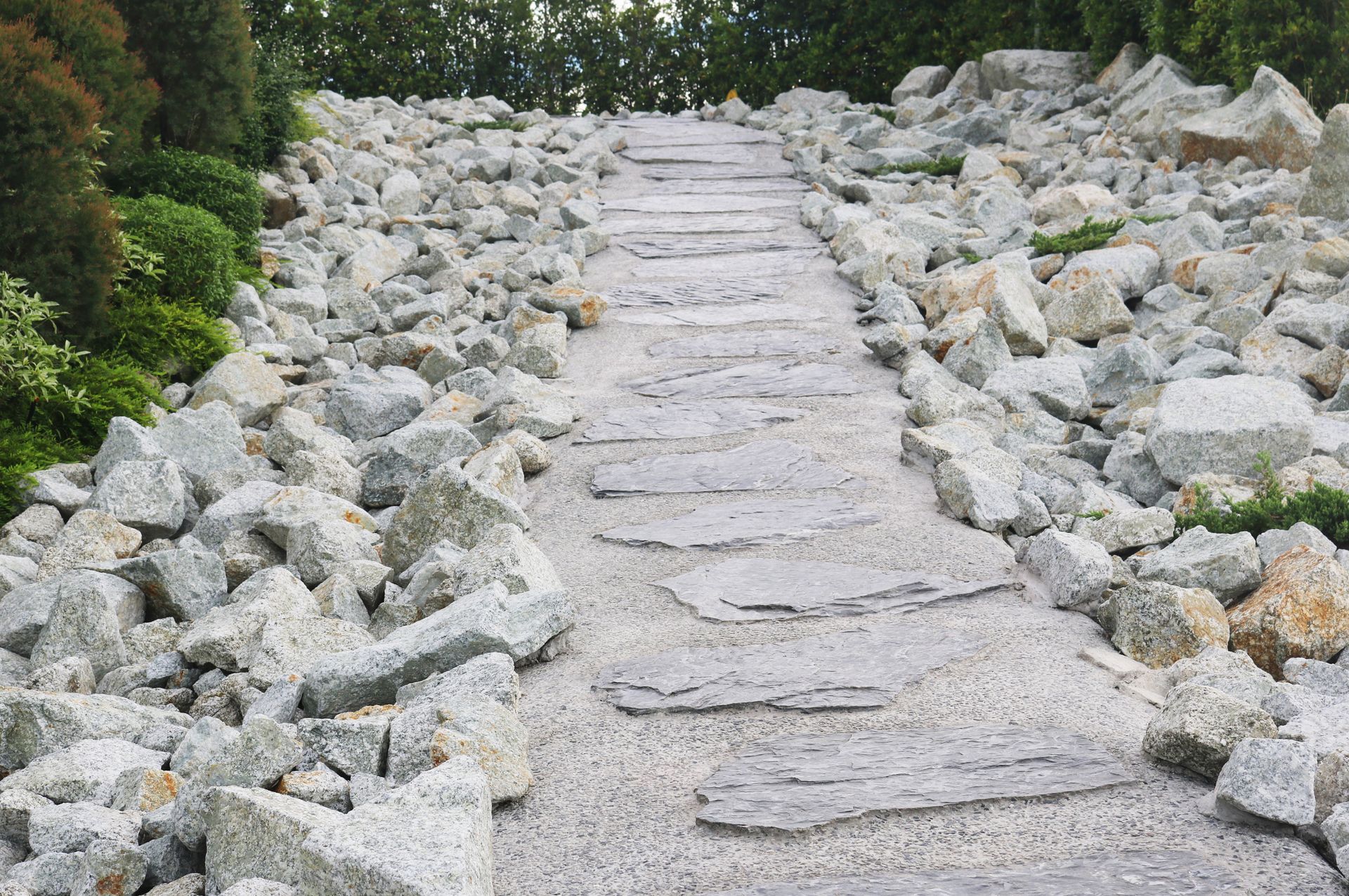 Stone pathway with stepping stones ascending through a rock garden, bordered by boulders and foliage.