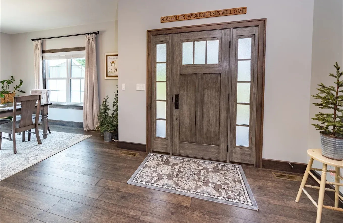 Entryway with wooden double doors and sidelights, rug, wooden floors, and a small tree.