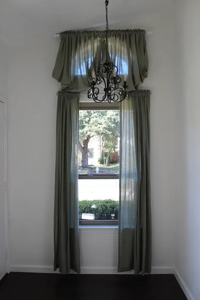 Tall window with olive green curtains, a chandelier, and a top valance.