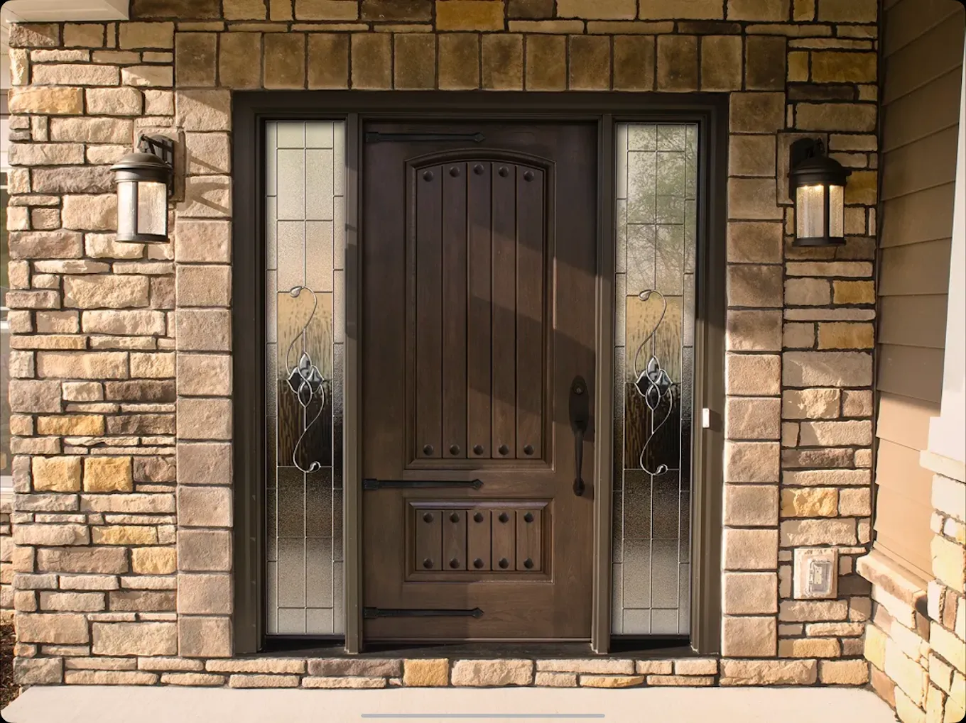 Brown wooden door with sidelights, set in stone facade, flanked by sconces.
