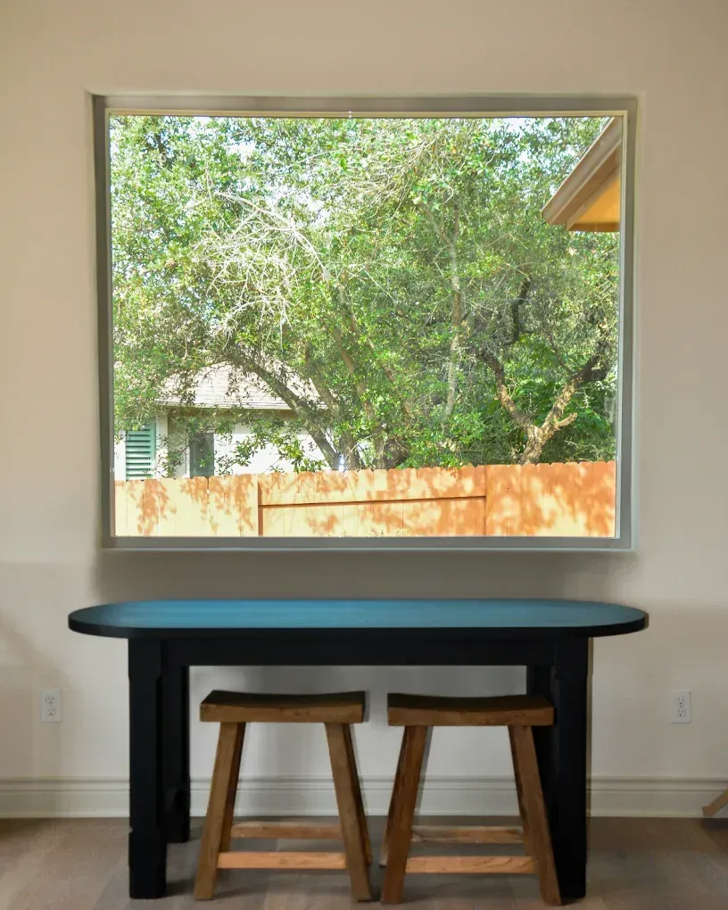 A black table with blue top and two wooden stools beneath a window overlooking trees and a fence.