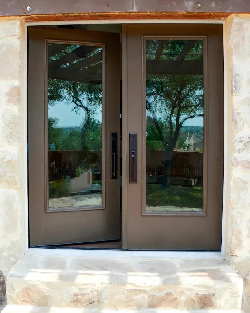 Double brown doors with glass panels, open to a sunny outdoor view, set in a stone wall.