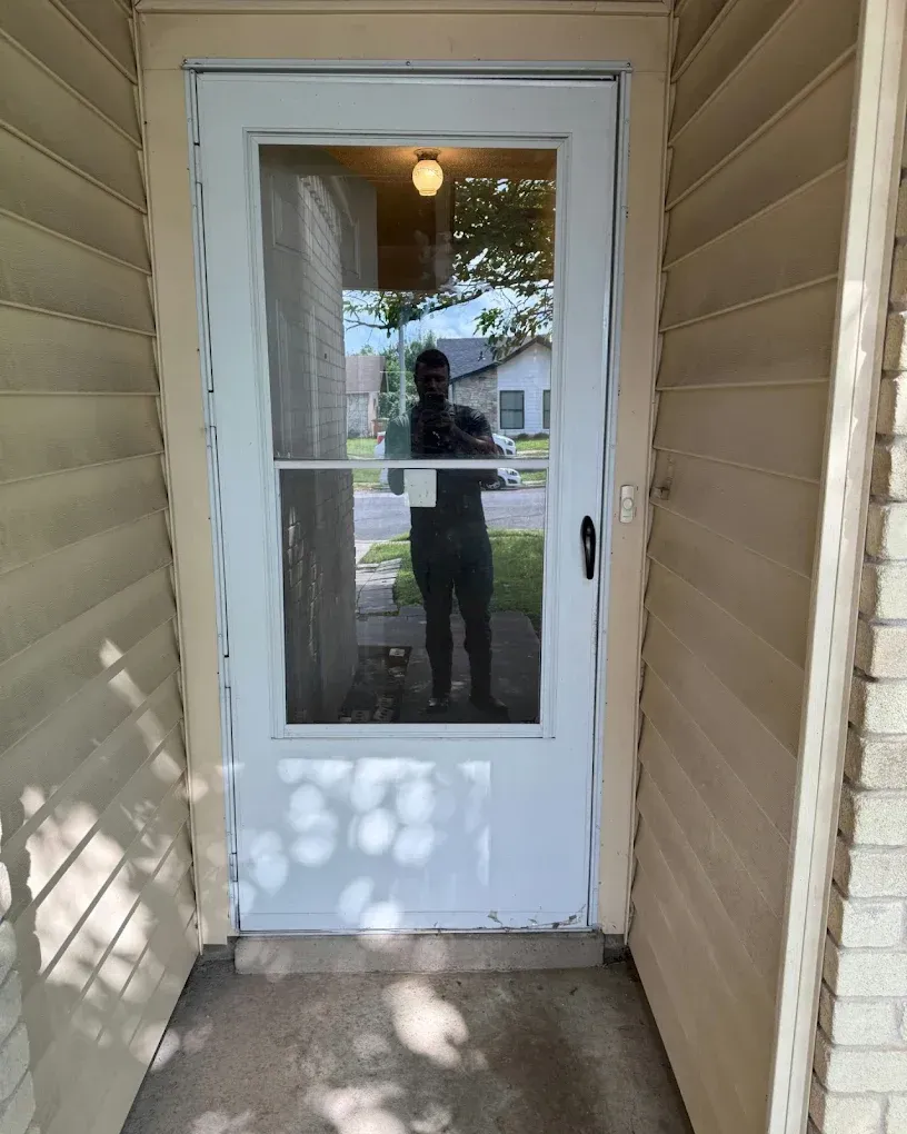White door with screen, tan siding, person reflected in the glass, front porch.