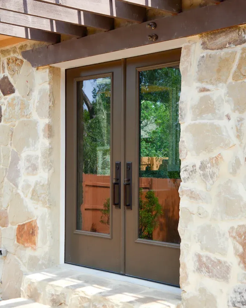 Double brown doors with glass panels set in a stone wall, beneath a wooden pergola.