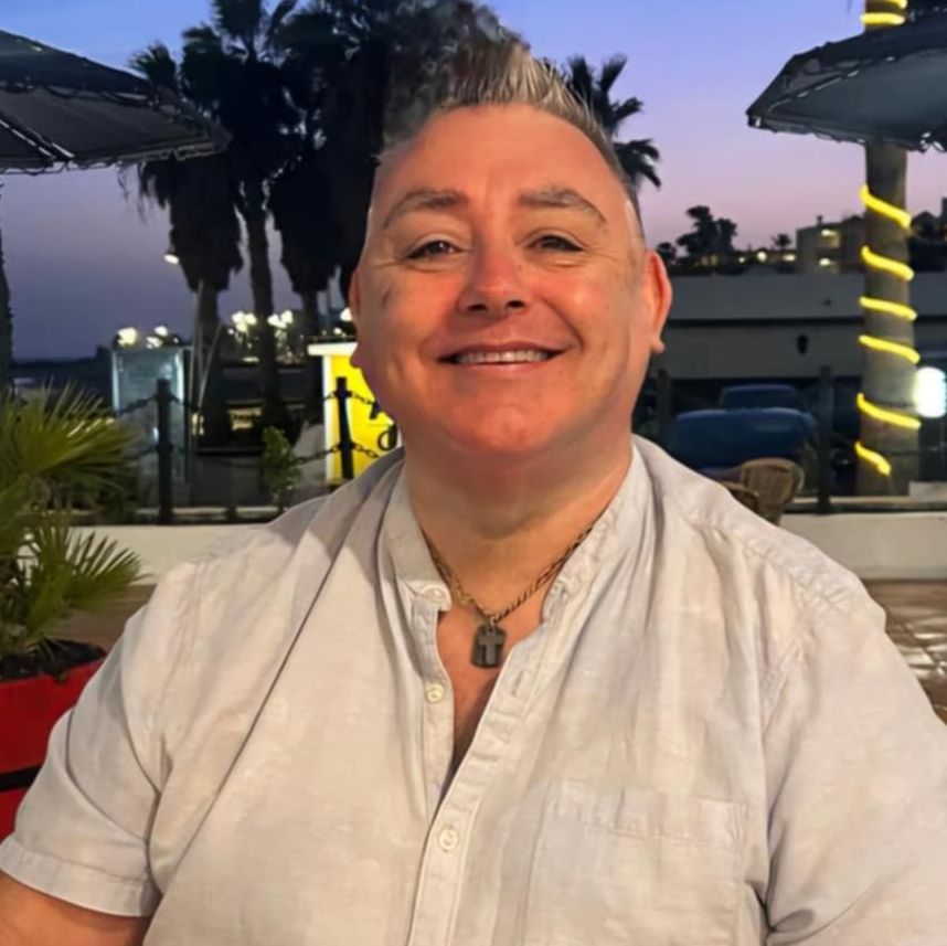 A man wearing a white shirt and a necklace smiles for the camera