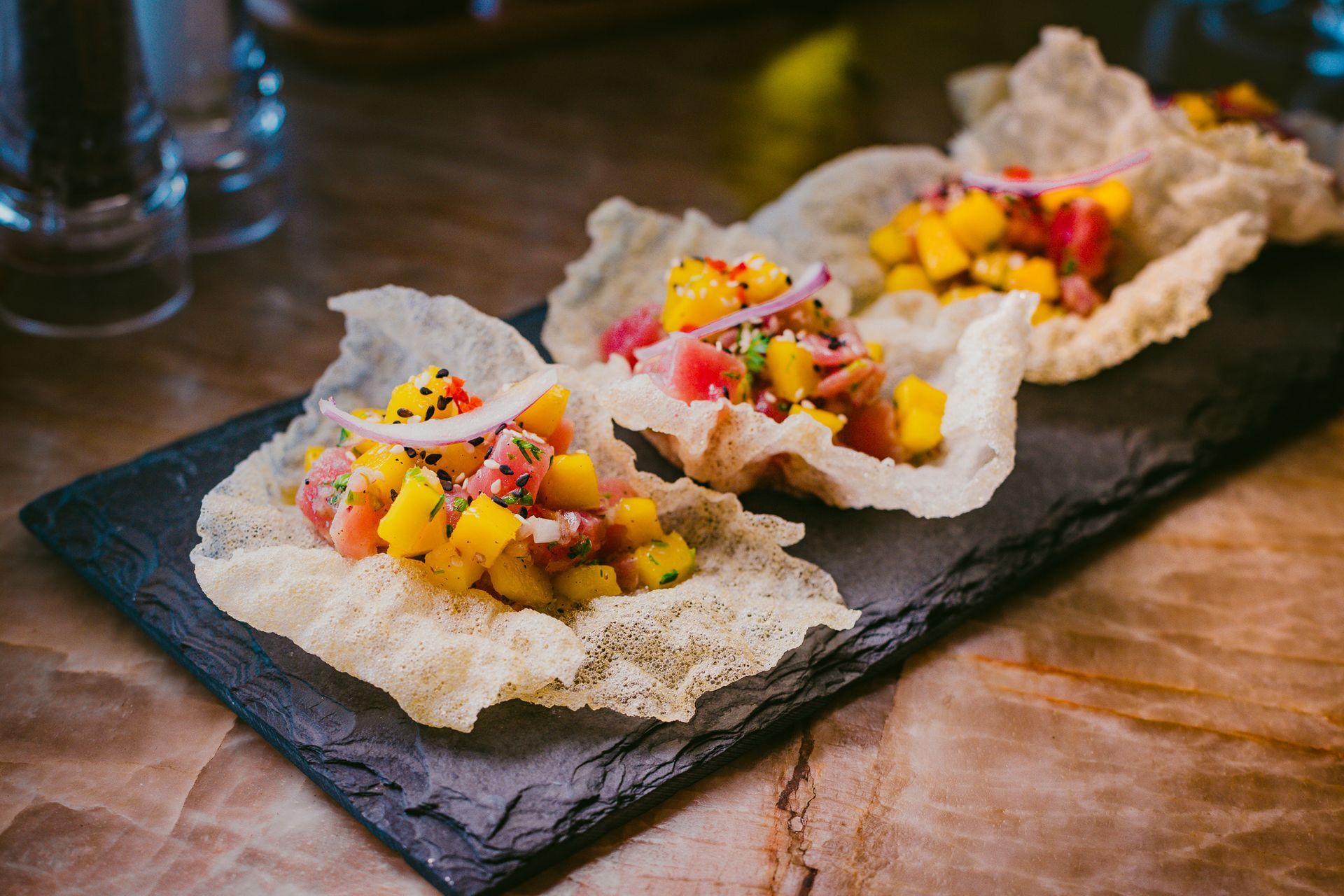 Tártaro de atum com manga servido em biscoitos de arroz crocantes, dispostos sobre uma tábua de ardósia.