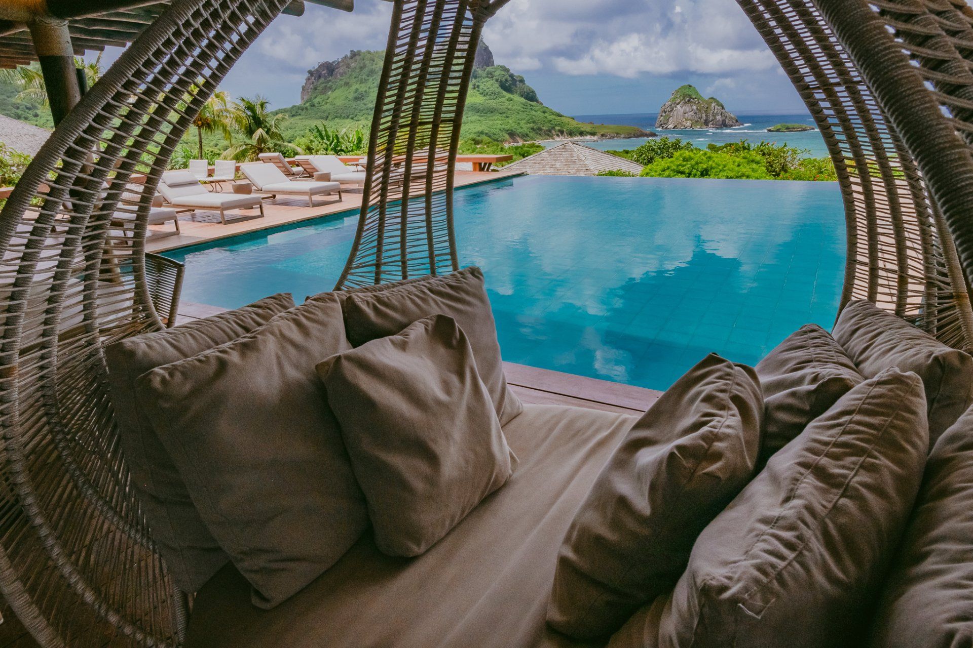 Lounge de vime com almofadas e vista para uma piscina azul de borda infinita e para o mar, com uma ilha ao fundo.