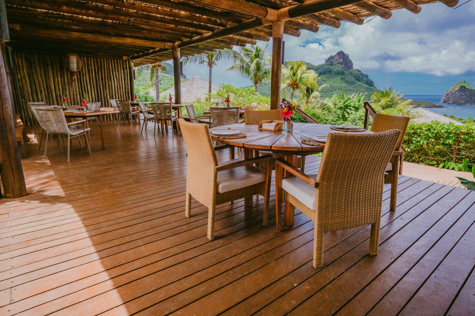 Restaurante ao ar livre com mesas/cadeiras de madeira, vista para o mar e muito verde.
