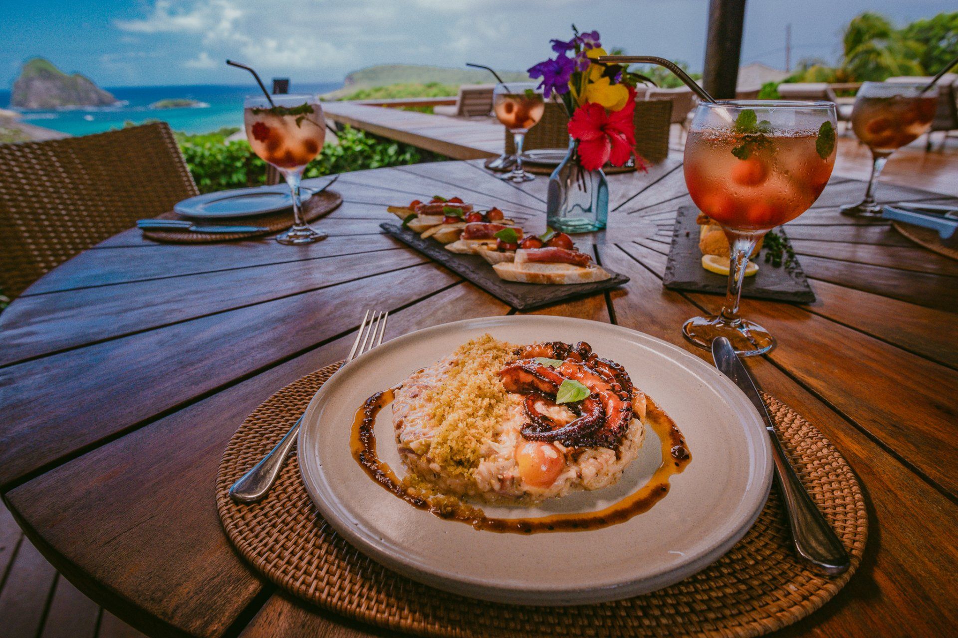 Refeição gourmet em uma mesa de madeira com vista para o oceano; coquetéis, aperitivos e prato principal presentes.