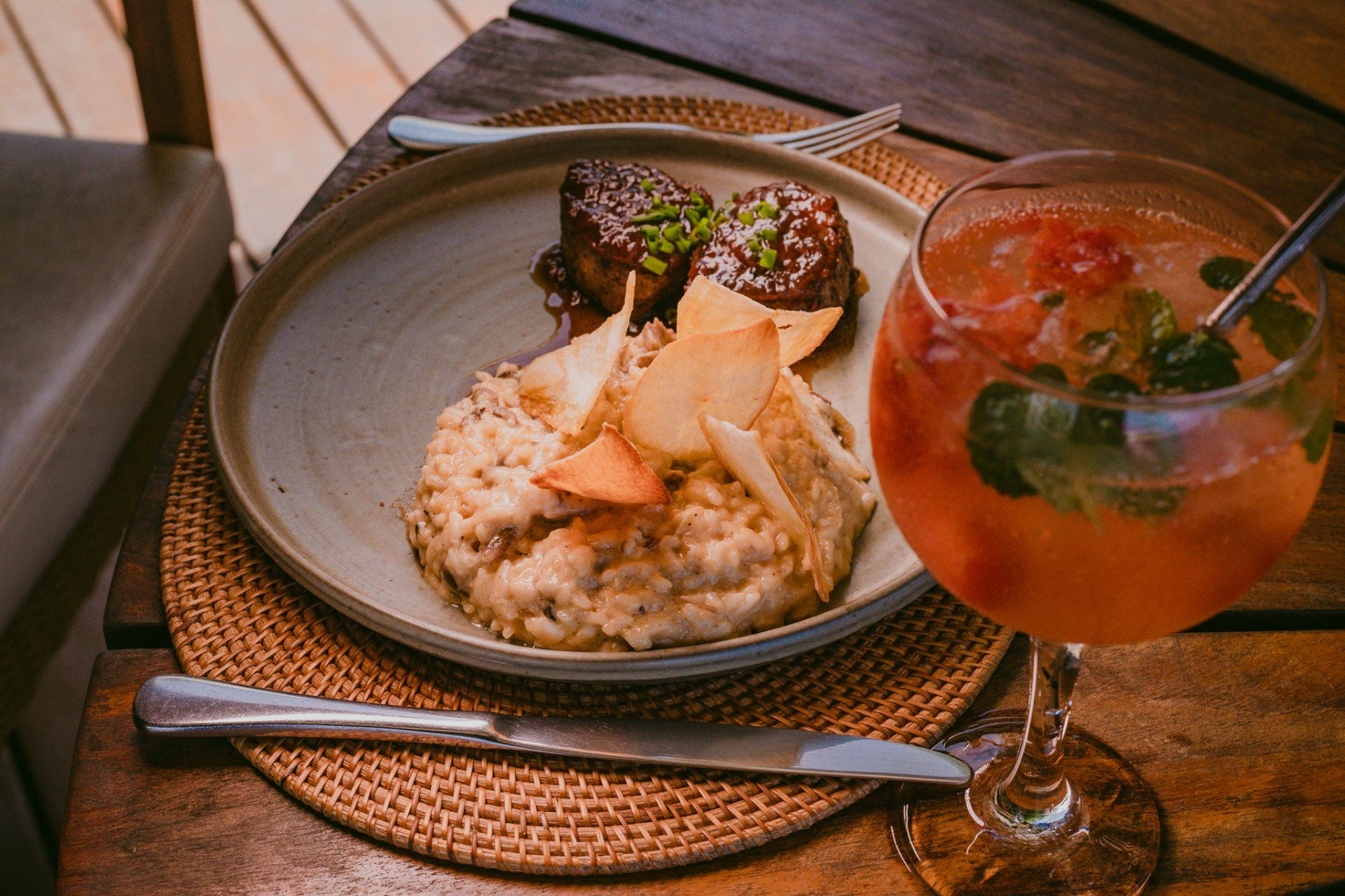 Prato de comida (risoto e almôndegas) com bebida, sobre uma mesa de madeira, perto de uma cadeira.