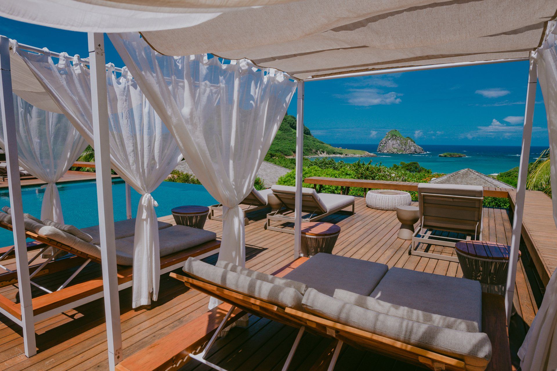 Espreguiçadeiras sob cortinas brancas perto de uma piscina com vista para o mar em um dia ensolarado.