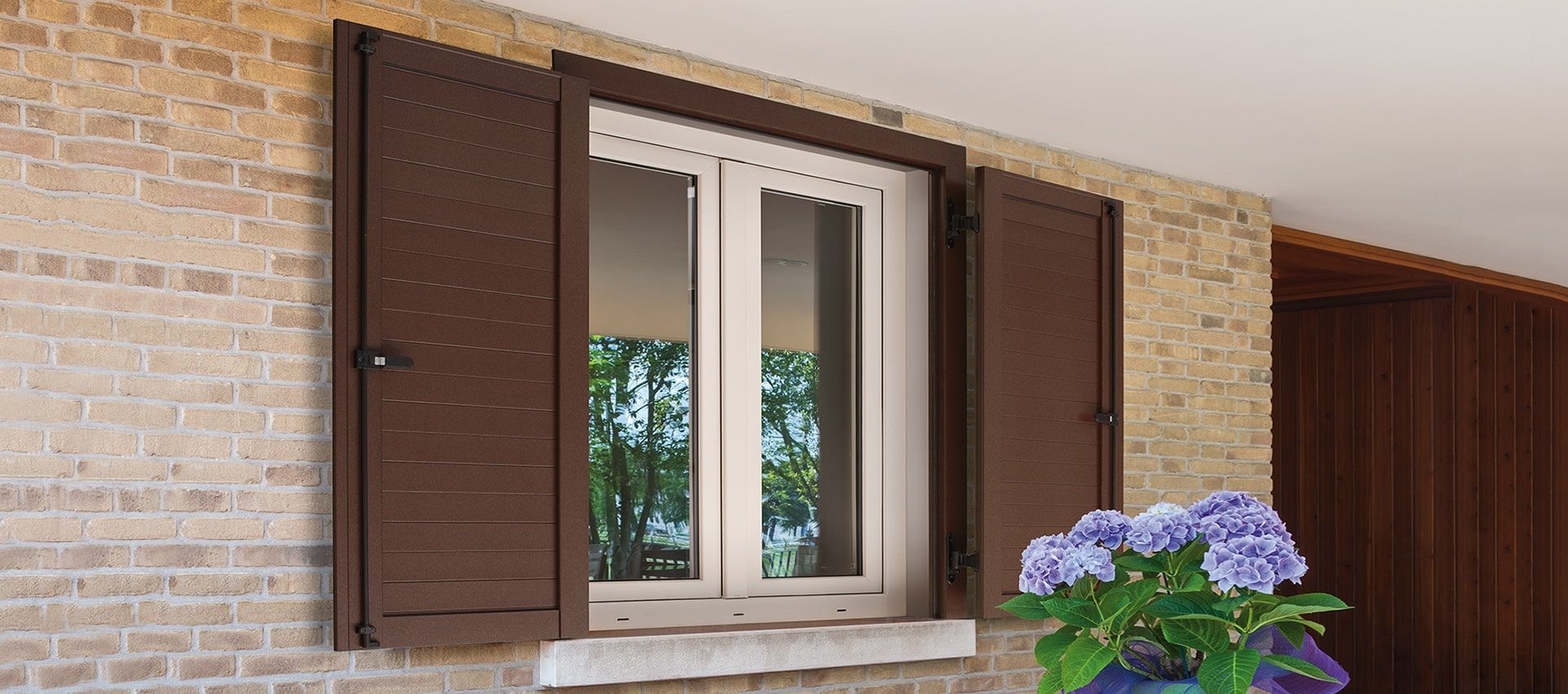 Una finestra con persiane marroni su un muro di mattoni accanto a un vaso di fiori.