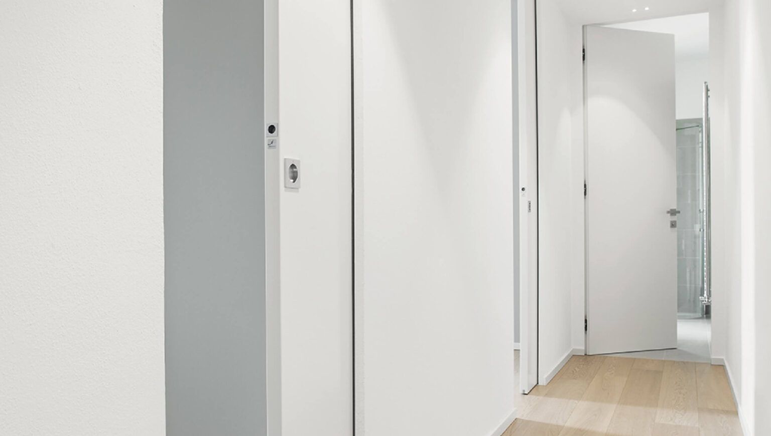 Un lungo corridoio con pareti bianche e pavimento in legno conduce ad un bagno.