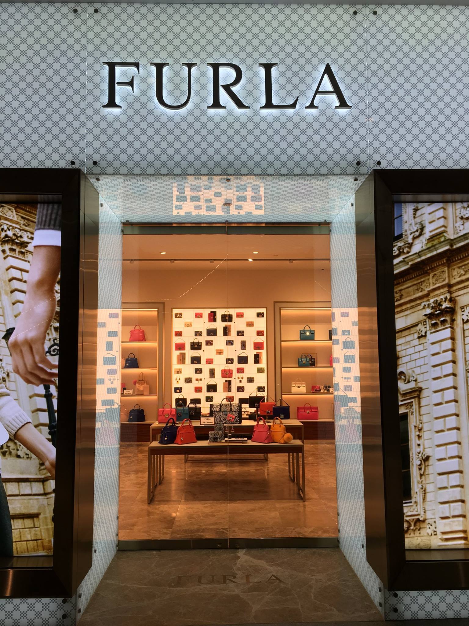 Furla storefront, showing bags in a brightly lit display. The shop's entrance is framed by marble panels with a logo overhead.