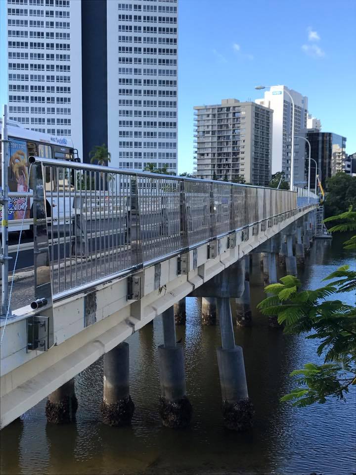 Bridge over water with a city skyline in the background. Gray and white buildings under a blue sky.