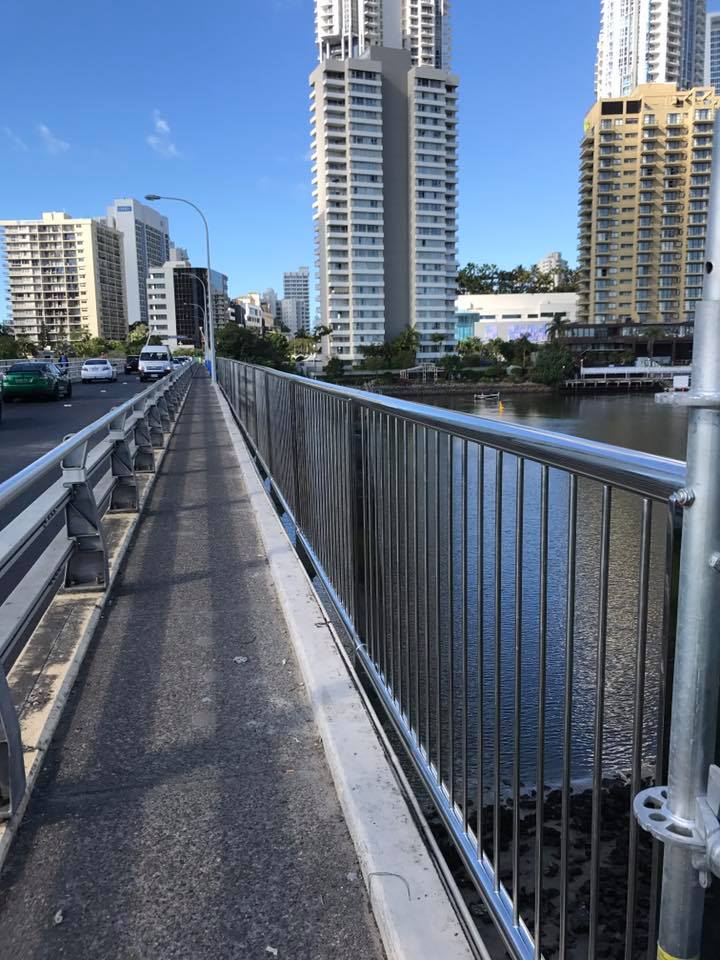 Bridge walkway with metal railings, city buildings in background, water on the right.