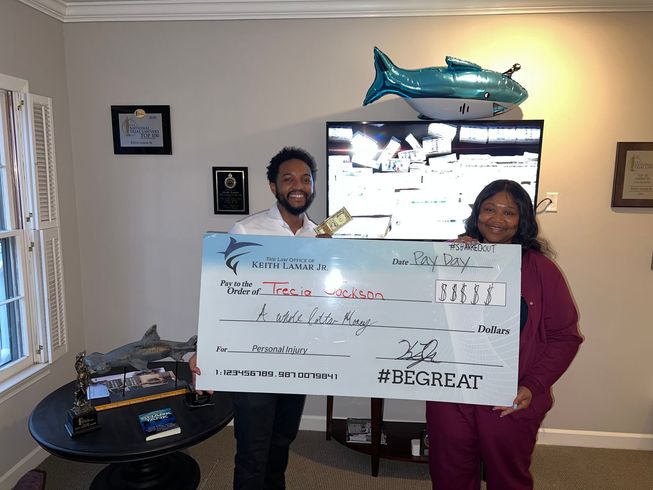 Two people holding a large check, smiling, indoors. A TV and shark balloon are visible.