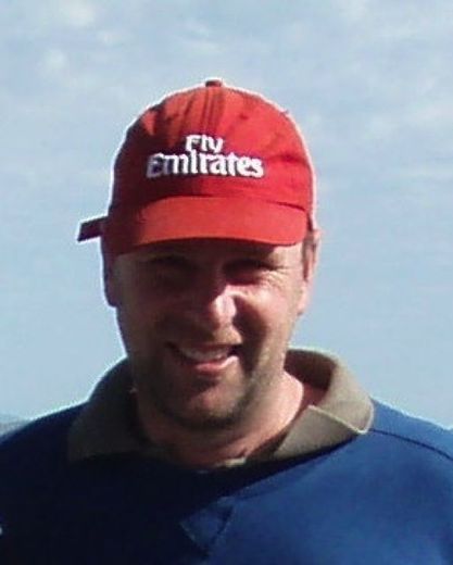 Man wearing a red Emirates cap and blue shirt, smiling outdoors against a cloudy sky