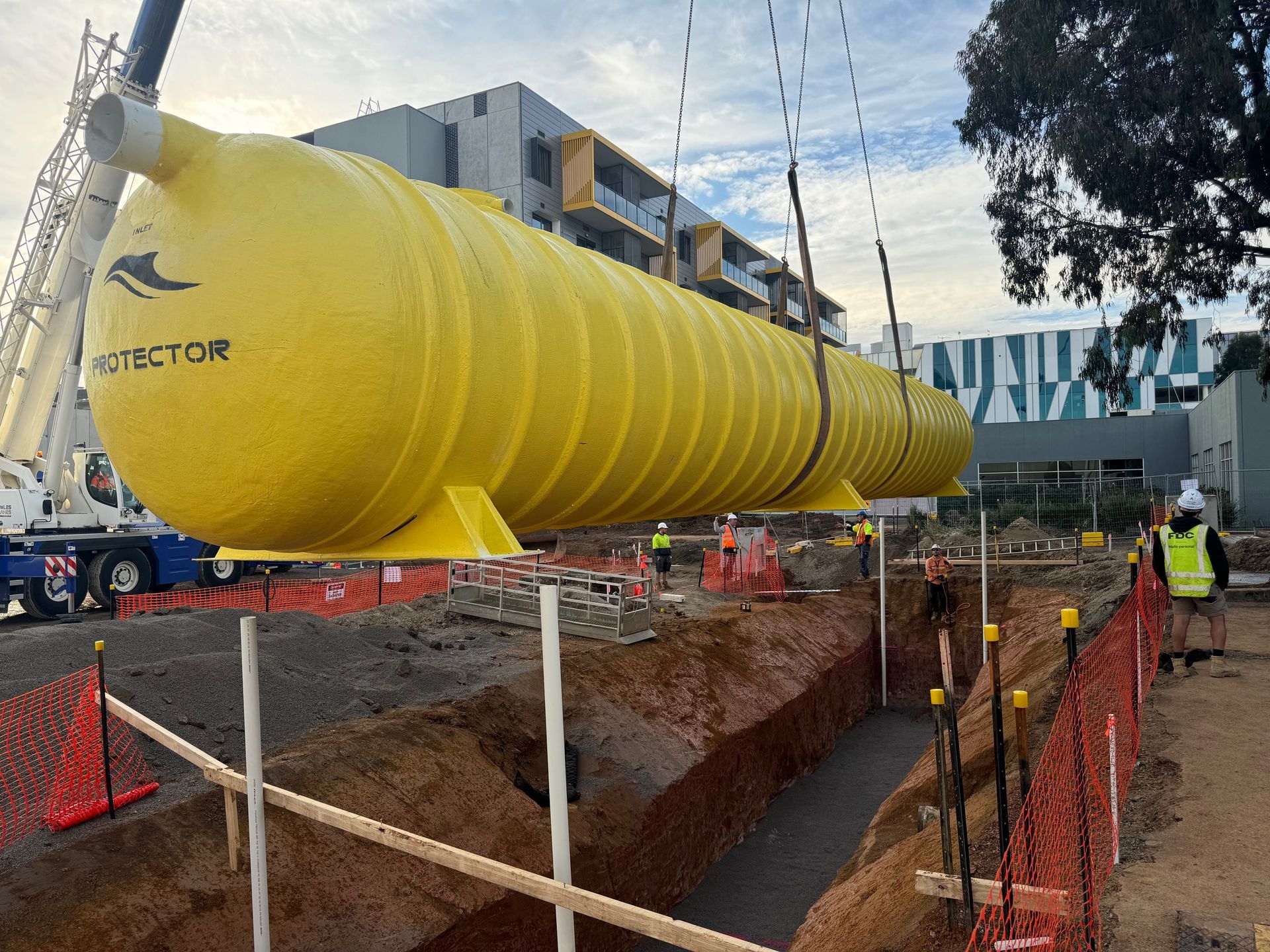 Yellow cylindrical water tank suspended by crane, being lowered into a prepared trench at a construction site.