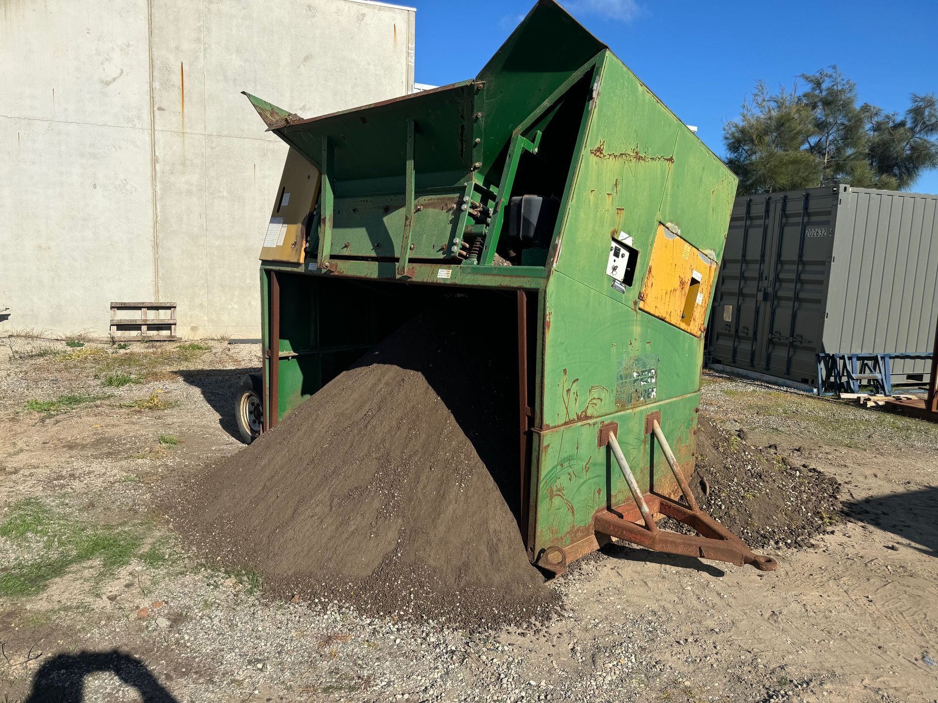 Green industrial soil sifter, tilted, with a pile of dark soil in front. Outdoors.