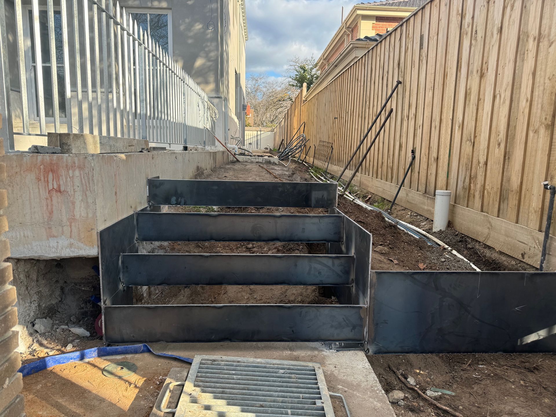 Metal stairs lead to a narrow dirt path between a building and wooden fence.