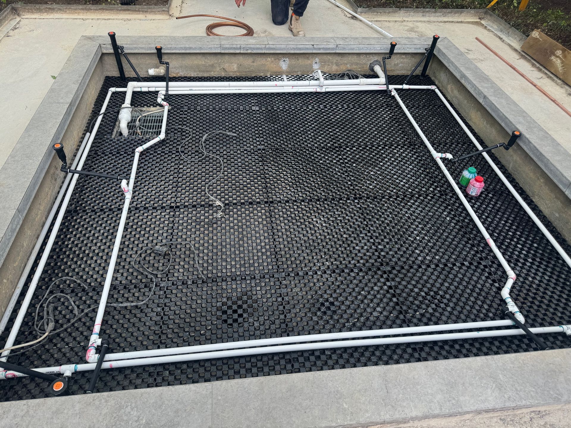 Black grid structure with white pipes in a concrete well. Electrical components and plumbing visible.