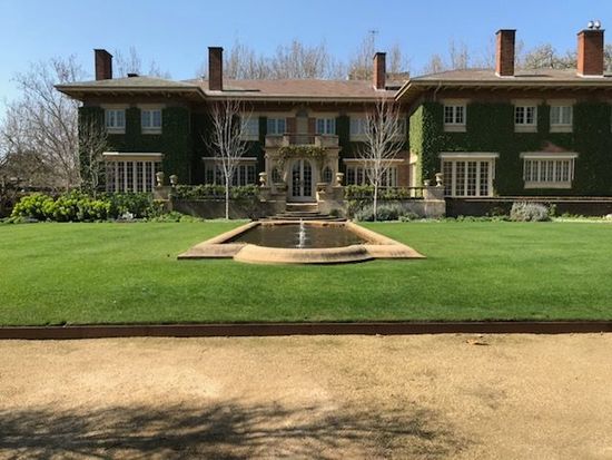 Large house with ivy-covered facade, fountain, and manicured lawn on a sunny day.