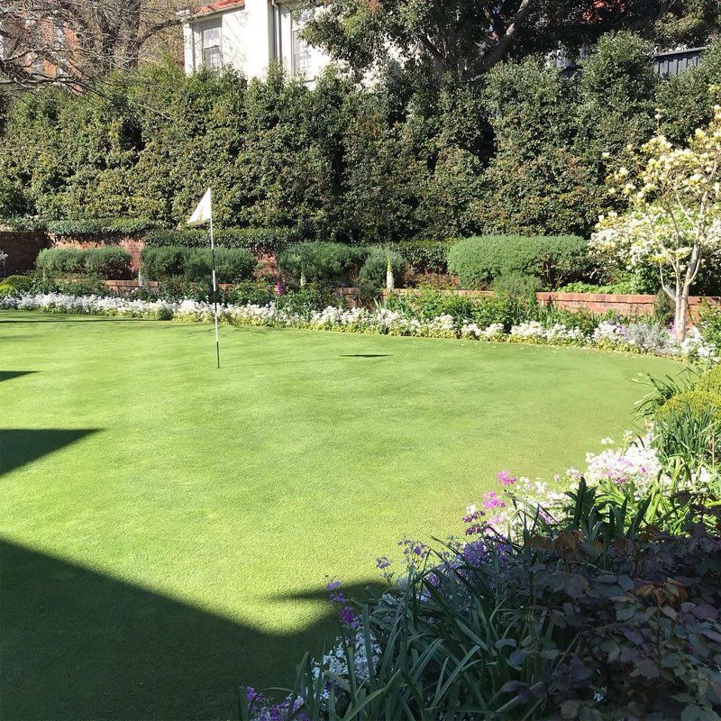 Lush green lawn with a golf flag, surrounded by flowerbeds and hedges, sunny day.